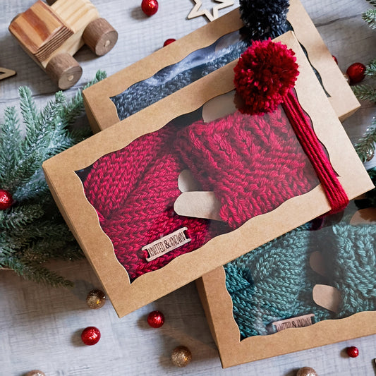 Red knitted fingerless gloves and an ear warmer in a gift box with a clear top on a festive background with Christmas decorations.