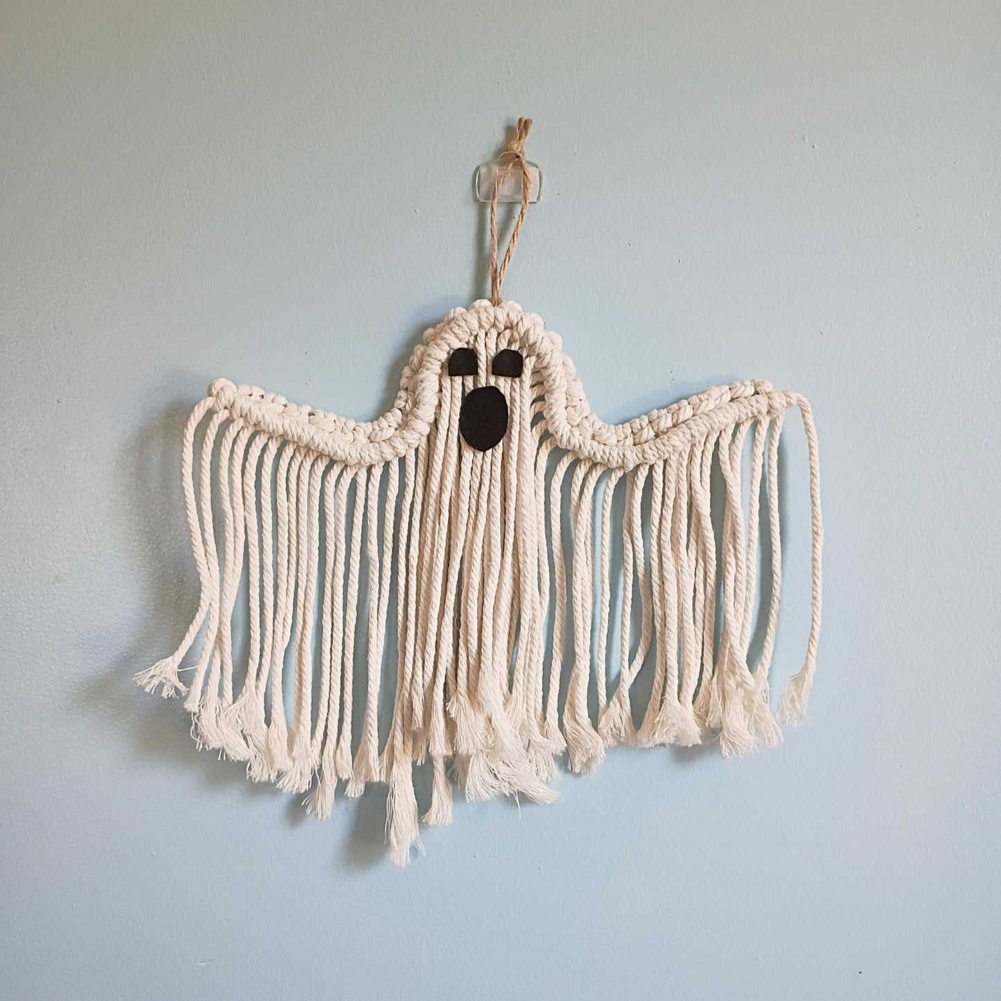 Handmade macrame ghost wall hanging in natural cotton rope with dark felt eyes and open mouth, hung on a light blue wall