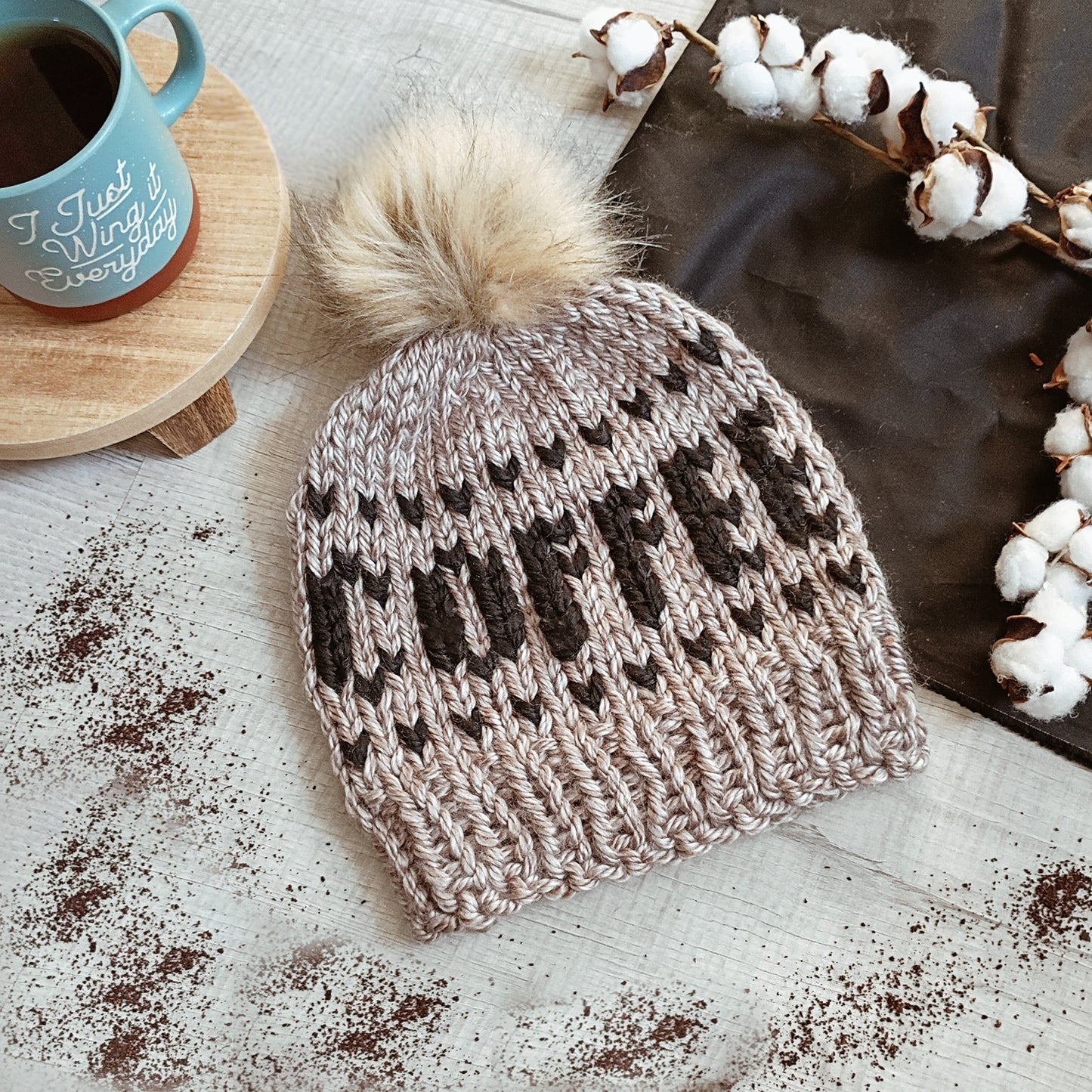 Knitted beanie with a pom-pom with the word coffee stitched on the front on a surface with coffee and cotton.