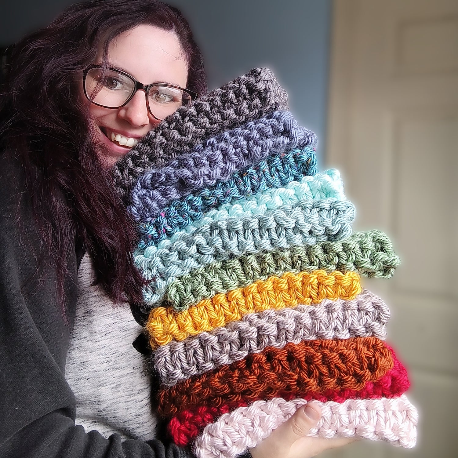 a women holding a stack of knitted neck cowls in colorful rainbow order starting with charcoal, dark blue, light blue, green, mustard, tan, burnt orange, red and pink 